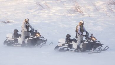 Two soldiers are seen operating two black-and-white Lynx Brutal Over Snow Reconnaissance Vehicles in the middle of a snowfield. The soldiers are wearing white as camouflage to blend in well with their surroundings.