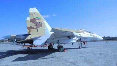 Two Sukhoi Su-30 planes are seen stationed on what appears to be a military air base. The planes are painted white, except for its camouflaged tails. The plane in front has "2401" painted on its tail, as well as the flag of Ethiopia. The background is a clear blue sky.