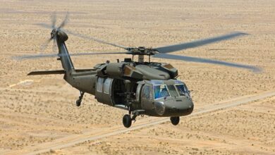 A Sikorsky UH-60M Black Hawk helicopter is seen flying over a desert plains. The dry, almost barren ground below has a pale yellow color. The helicopter's four spinning blades are blurry from its rotation. The helicopter's paint is dark green.