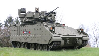 A Bradley M2A4E1 Fighting Vehicle is seen stationed at a grassy area with thin trees in the back. The vehicle is a tracked armored unit that is painted grayish green. Its turret points northeast, and its body is facing to the right side of the image. Its model name is painted in white on its right side, with a gray and red background.