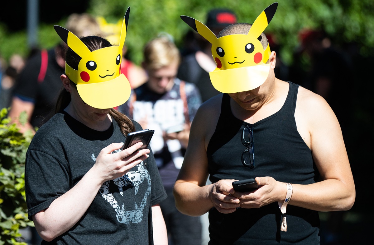 Two German adults wearing Pikachu sun visors play Pokemon GO on their phones in a park