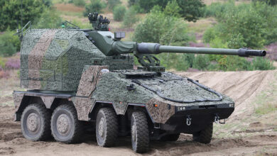 A KNDS Remote Controlled Howitzer 155 mm is seen on a grassy and sandy area. The eight-wheeled vehicle is painted with textured green and brown camouflage. Its turret is jutting forwards from the back. In the ground, trees and bushes grow scattered.