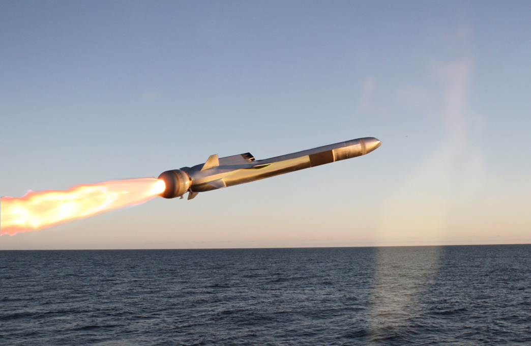 Kongsberg's Naval Strike Missile (NSM) is seen front and center, launching from the left. A jet of red-orange flame streaks from its back. The background is a deep blue ocean, with a cloudless sky farther back.