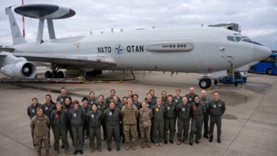 The women of the NATO Airborne Early Warning and Control Force pose for a photo in front of a NATO AWACS plane. They appear to be on a plane runway. The soldiers are all wearing green fatigue overalls. The sky in the background is covered by white heavy clouds.