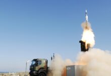 A SAMP/T air defense system is seen firing off a ballistic missile from the back of the military truck. Smoke and bright white-red fire can be seen jutting out of the missile as it flies straight upward. The background is a clear blue sky.