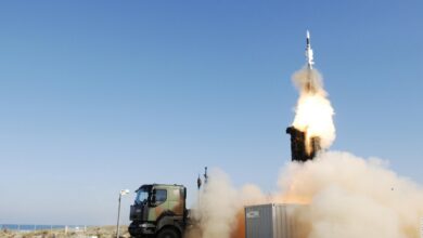 A SAMP/T air defense system is seen firing off a ballistic missile from the back of the military truck. Smoke and bright white-red fire can be seen jutting out of the missile as it flies straight upward. The background is a clear blue sky.