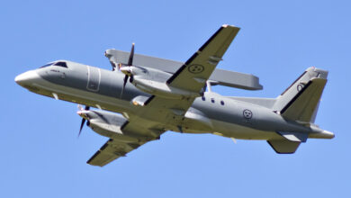 A Saab 340 aerial early warning and control plane is seen flying in clear blue skies. Its long, rectangular radar system is seen connected and propped up parallel to its body. The aircraft is painted gray. The photo was taken underneath the plane.