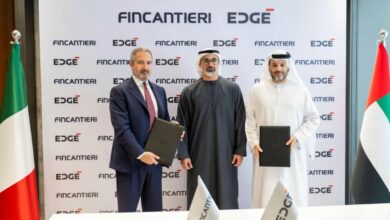 Fincantieri CEO and Managing Director Pierroberto Folgiero, Abu Dhabi crown prince Khaled bin Mohamed Al Nahyan, and EDGE Group chairman Faisal Al Bannai stand to take a photo op during a signing ceremony. Folgiero and Al Bannai hold leather dosiers, which may be holding the signed documents. Italy's flag is seen displayed on the left, and UAE's is on the right. On a white table in front of them, two flags printed with Fincantieri and EDGE's logos are also propped up. Behind them is a white screen with the companies' logos printed in a repeating pattern.