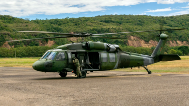 A Colombian Army Black Hawk helicopter is seen grounded in a large open cemented field. The helicopter is painted various hues of dark green as camouflage. A soldier in camouflage is seen standing by the cockpit. "EJERCITO DE COLOMBIA" is seen painted in black on the aircraft's tail boom. The background is a mountain covered by a lush green forest, and a bright blue sky with big white clouds.