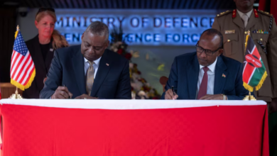 Secretary of Defense Lloyd J. Austin III (left) and Kenyan Defense Minister Aden Bare Duale (right) sit beside each other while signing the papers of the five-year framework for defense cooperation. They are flanked by one military personnel each on the background. A US flag is placed on Austin's side, while a Kenyan flag is on Duale's.